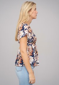 Bloemige blouse in marineblauw met roze en oranje bloemen. Korte mouwen, gathering in de taille en een relaxed fit. Gecombineerd met lichtblauwe jeans.