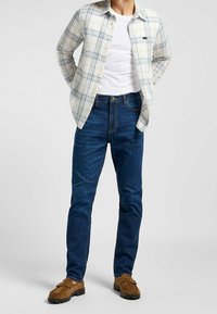 Camisa de cuadros claros abotonada, camiseta blanca, vaqueros azul oscuro, mocasines de ante marrón. La camisa tiene cuello y mangas largas; los vaqueros son ajustados.