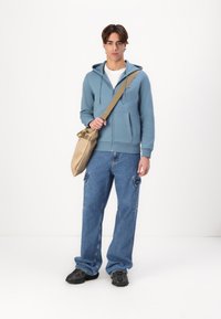 Haut zippé bleu sur une chemise blanche, pantalon cargo en denim, baskets noires et un sac à épaule beige avec un haut plié.
