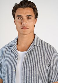 Koszula z krótkim rękawem zapinana na guziki w jasnoniebiesko-biały wzór w paski, wykonana z fakturowanej tkaniny. Posiada kołnierz z wycięciem i guziki z przodu.