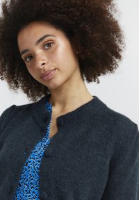 Mørkegrå cardigan med rund halsudskæring og knaplukning, båret over en blå og sort mønstret bluse med en glat tekstur.