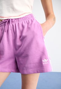 Lilla treningsshorts med elastisk livlinje og snøring. Har hvite sidestreker og et brodert Adidas-logo på kanten.