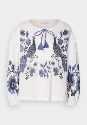 White cotton blouse with blue floral and peacock embroidery, featuring a round neckline, gathered long sleeves, and a tie detail at the front.
