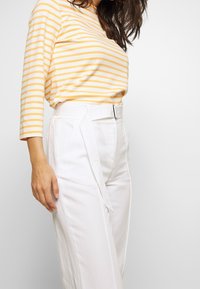 Striped yellow and white long-sleeve top with white high-waisted trousers featuring a belt detail and side pockets. Minimalist design.