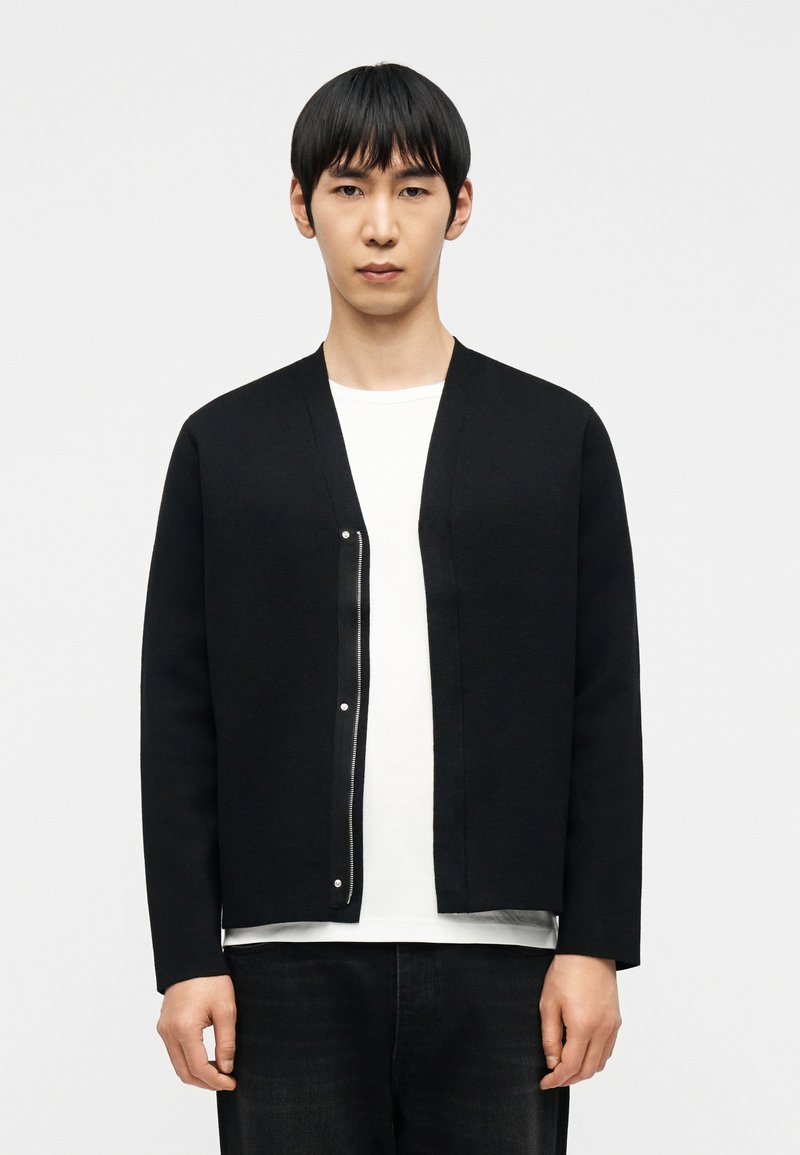 Νέος άνδρας που φοράει μαύρη ζακέτα με φερμουάρ  πάνω από λευκό πουκάμισο και μαύρο παντελόνι, στέκεται μπροστά σε μια απλή φωτεινή επιφάνεια.