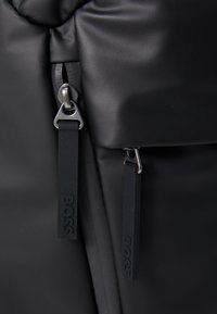 Sac à dos en matériau texturé noir avec des tirettes de fermeture éclair doubles et des accents en caoutchouc inscrits "BOSS". Design compact avec des lignes épurées et une fermeture sécurisée.