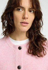 Cardigan rose clair en tissu texturé, avec un col rond et trois boutons noirs à l'avant, porté sur un haut blanc.