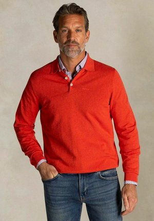 Middelbare man met baard die een rood polo met lange mouwen draagt over een patroonhemd en een blauwe spijkerbroek, staand met één hand in zijn zak.