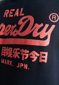 Textil negro con un gran logo texturizado en rojo que dice "REAL SuperDry" con caracteres japoneses debajo y "MARK. JPN." en la parte inferior.