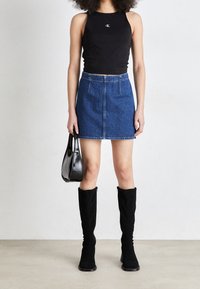 Kvinna i svart ärmlös topp, blå denim minikjol, svarta knähöga stövlar, håller en svart handväska mot en enfärgad bakgrund.