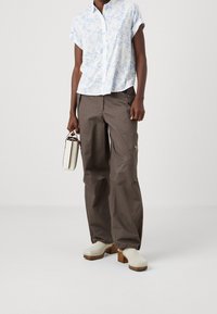 Weißes kurzärmliges Hemd mit blauer Blumenmuster, kombiniert mit braunen, weit geschnittenen Cargo-Hosen. Hält eine kleine weiße Tasche und trägt cremefarbene Clogs.