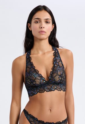 Donna con capelli lunghi e scuri che indossa un reggiseno in pizzo nero iridescente e mutandine abbinate, in piedi contro uno sfondo chiaro e uniforme.