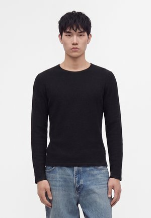 Jeune homme aux cheveux courts et noirs porte un pull noir uni à manches longues et un jean bleu clair, debout devant un fond blanc.