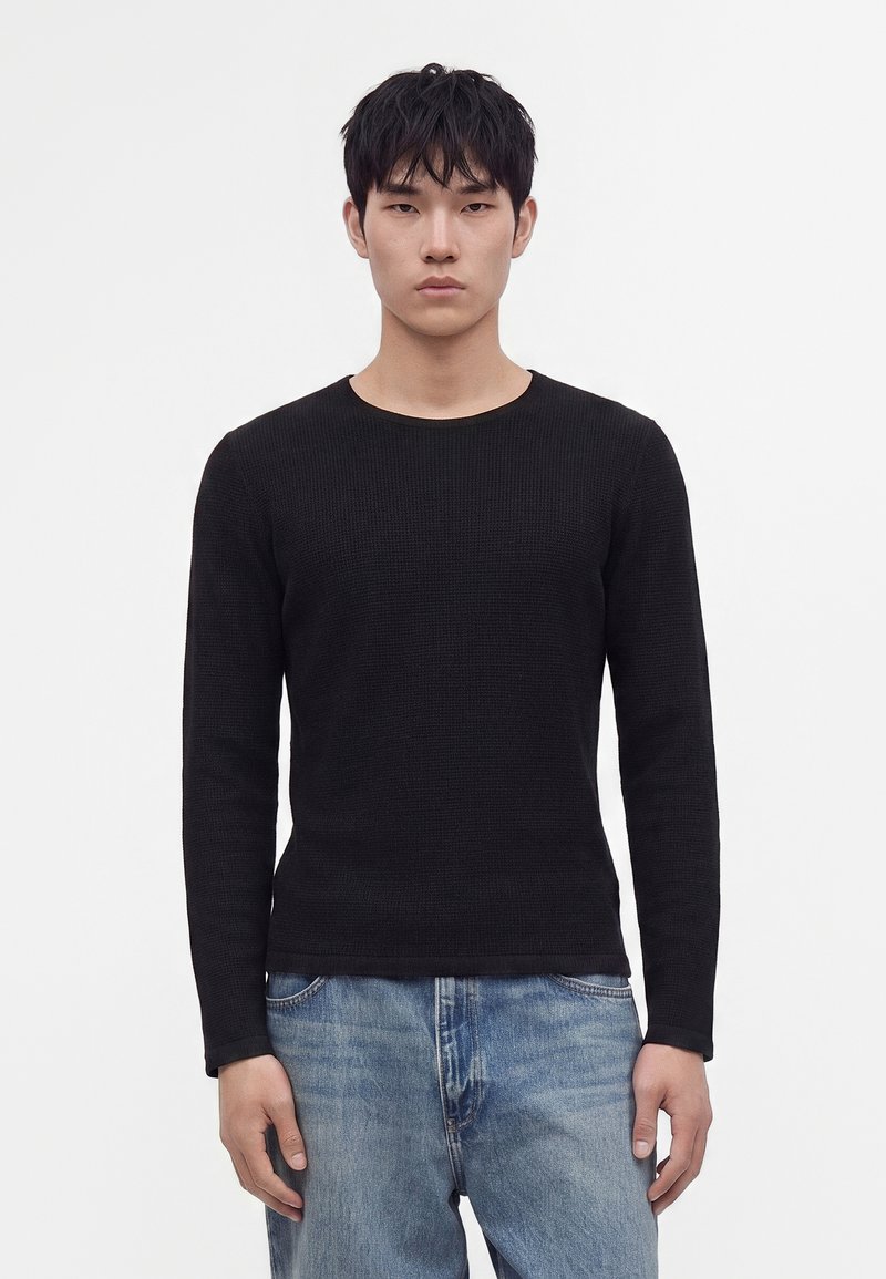 Jeune homme aux cheveux courts et noirs porte un pull noir uni à manches longues et un jean bleu clair, debout devant un fond blanc.