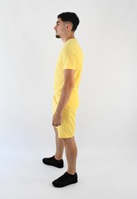 Camiseta de algodón amarilla combinada con pantalones cortos a juego, ambos ajustados. Llevado con zapatillas negras, visto de lado contra un fondo neutral.