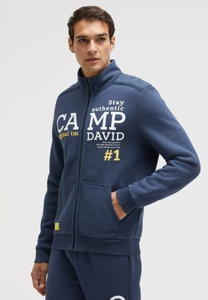 Junger Mann trägt eine dunkelblaue Jacke mit Reißverschluss und Hose mit der Aufschrift „Camp David“, steht mit einer Hand in der Tasche und schaut zur Seite.