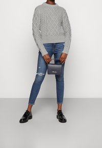 Osoba w szarej swetrze w warkocze, w podartych niebieskich rurkach, czarnych butach, trzymająca małą granatową torebkę na łańcuszku.