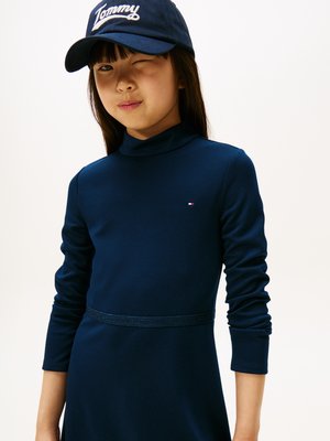 Marineblauwe lange jurk met hoge hals en een gestructureerde tailleband. Een marineblauwe pet met het witte "Tommy" logo maakt de outfit compleet.