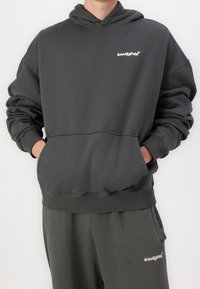 Sweat à capuche gris foncé avec une poche kangourou, des poignets et un ourlet côtelés. Présente un petit logo blanc sur la poitrine. Texture douce, semblable au coton.