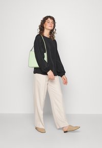 Blusa preta de mangas longas com tecido texturizado, calças beje de perna larga e sapatos slip-on de tecido bege. A segurar uma bolsa verde clara.
