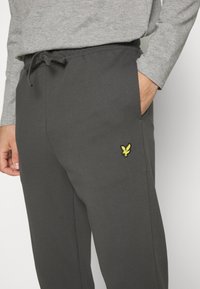 Donkergrijze sweatpants van zacht materiaal, met een elastische tailleband met koord en een kleine gele adelaar logo op de dij.