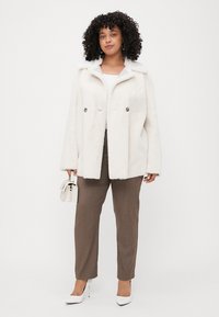 White faux fur coat with a collar, double-breasted style, paired with brown tailored pants and white stilettos, holding a small white bag.
