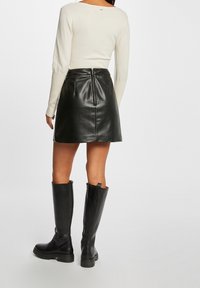 Kobieta ubrana w kremowy top z długim rękawem, czarną mini spódnicę ze skóry z tylnym zamkiem oraz czarne skórzane kozaki do kolan, stojąca tyłem.