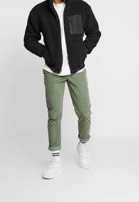 Zwarte fleece jack met een voorkant ritszak, gecombineerd met lichtgroene broek, witte sneakers en een wit T-shirt, wat een casual stijl laat zien.