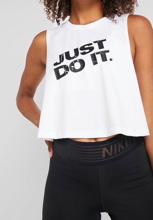 Femme portant un crop top sans manches blanc avec le texte "JUST DO IT." et des leggings noirs taille haute avec une ceinture logo Nike.