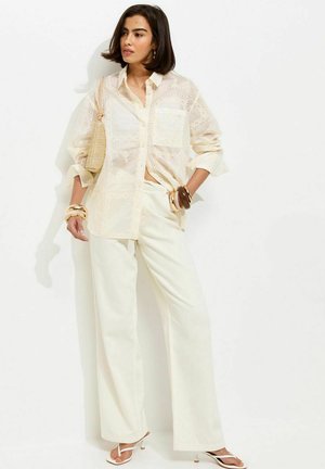 Vrouw in loshangend wit geborduurd overhemd en broek met wijde pijpen, met een gevlochten tas, armbanden en witte sandalen met hak, staand tegen een witte muur.