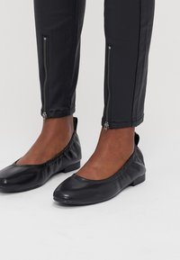 Zwarte leren platte schoenen met een afgeronde neus, elastisch en met een lage hak. Zwarte broek met zijritsen op de enkel.