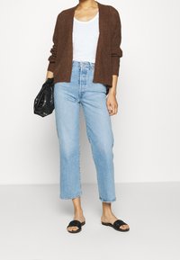 Hnědý pletený kardigan, bílý trikoto, modré džíny s vysokým pasem a černé pantofle. Vyznačuje se uvolněným střihem a hladkou texturou.