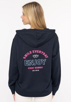 Dunkelblaues Hoodie mit einer Fronttasche und einer Kapuze mit Kordelzug, auf der Rückseite mit mehrfarbigem Text: "LACHE JEDEN TAG, GENIEßE JEDEN MOMENT, GEGRÜNDET 2014."