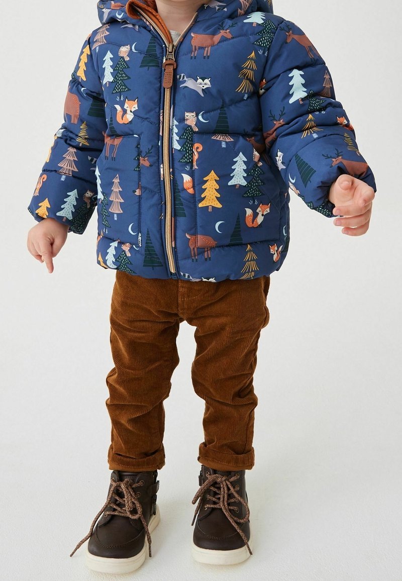 Tout-petit portant une doudoune bleue avec un imprimé d’animaux de la forêt, un pantalon en velours côtelé marron et des bottes marron foncé avec des semelles beiges.