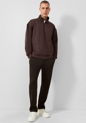 Man draagt een donkerbruine quarter-zip trui, zwarte broek en witte sneakers, staande tegen een effen lichtgrijze achtergrond.