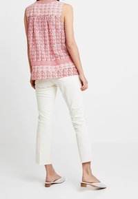 Top senza maniche rosso con motivi geometrici bianchi, abbinato a pantaloni avorio. La persona indossa scarpe bianche senza lacci con tacco basso.
