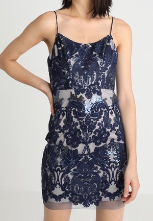 Mujer con un vestido mini sin mangas con superposición de encaje floral azul marino y tirantes finos sobre un fondo claro.