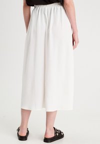 Culottes blanches fluides en tissu texturé, avec une taille élastique et des poches latérales. Portées avec des sandales à glissière noires ornées.