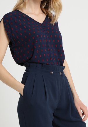 Frau trägt eine marineblaue, ärmellose Bluse mit kleinen roten Mustern, die in eine hoch taillierte marineblaue Hose mit vorderen Knöpfen gesteckt ist.