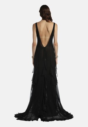 Hosszú barna hajú nő fekete, ujjatlan ruhában, mély V-alakú hátkivágással és réteges fodros uszállyal, sima háttér előtt állva.