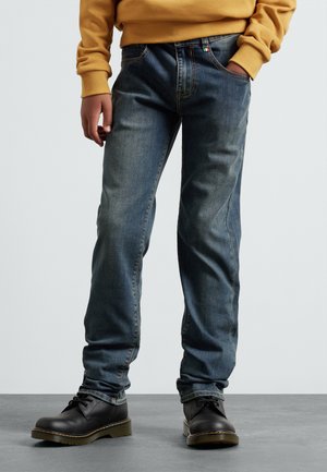 Jean en denim bleu avec une coupe droite, dotés de cinq poches et d'un surfil contrasté. Portés avec des bottes en cuir noir et un sweat-shirt moutarde.