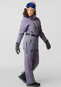 Paarse geïsoleerde skibroek met meerdere ritszakken, een riem en een hoge kraag, gecombineerd met grijze gloves en een roze beanie en goggles.