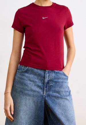 Mujer con camiseta ajustada Nike de color rojo oscuro y pantalones vaqueros azules sueltos, con una mano en el bolsillo, posando frente a un fondo liso.