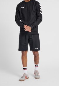 Hombre con sudadera negra de hummel y pantalones cortos, calcetines blancos con rayas negras y rojas, y zapatillas grises con acentos rojos, de pie con las manos entrelazadas.