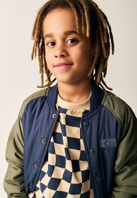 Marineblå og olivengrøn bomberjakke med trykknapper, båret over en beige og marineblå ternet t-shirt. Glat stof og afslappet pasform.