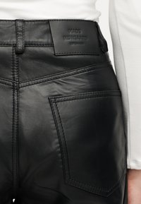 Detailný záber na čierne kožené nohavice s zadným vreckom a prišitým štítkom s nápisom "Mads Nørgaard Copenhagen", nosené s bielym topom.