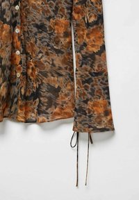 Chemise à manches longues avec un motif floral orange et brun, dotée de liens au niveau des poignets et de boutons blancs sur le devant.
