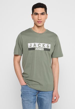 Giovane uomo con capelli corti e ricci che indossa una maglietta verde "JACK & JONES" e jeans blu, in piedi con una mano in tasca.