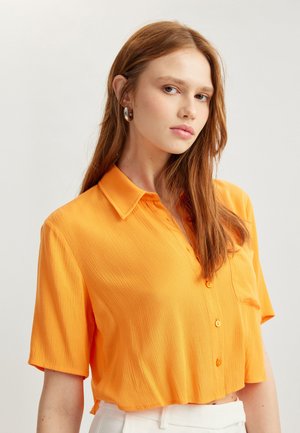 Kortærmet, krave skjorte i lys orange stof. Har en struktureret overflade, knaplukning foran og en brystlomme. Croppet design.