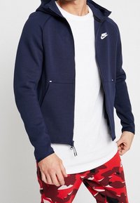 Marinblå zip-up hoodie med en framficka, vit Nike-logga på bröstet och en huva. Bärs över en vit t-shirt och röda kamouflagebyxor.